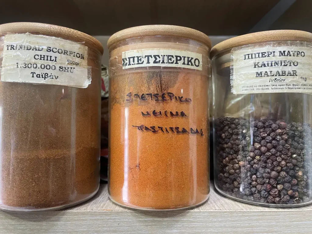 Glass jars of Spetseriko spice blend, Trinidad Scorpion chili, and smoked Malabar black pepper inside a traditional Corfu spice shop.